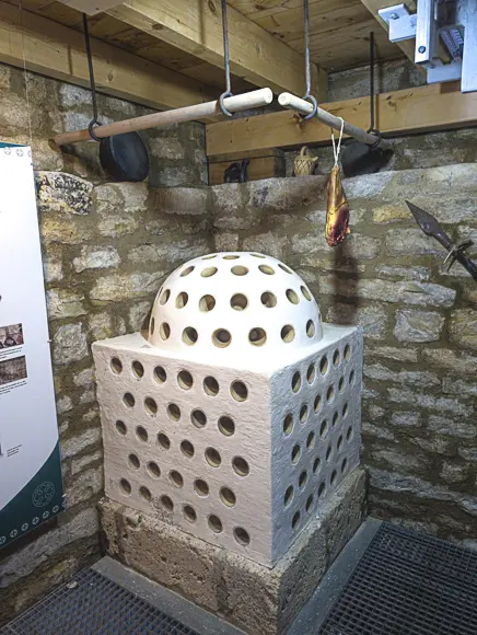 Ein Ausstellungsstück, das einen mittelalterlichen Kachelofen oder Rauchofen in einer rustikalen Umgebung mit Steinmauern und Holzbalken darstellt. Der Ofen besteht aus einem quadratischen Sockel und einer gewölbten Haube, beide sind aus hellem Material gefertigt und mit zahlreichen runden Löchern perforiert, vermutlich zur Wärmeabgabe. Über dem Ofen hängen von Holzbalken herab ein Kochtopf und ein geräuchertes Stück Fleisch (Schinken) an einer Schnur, was auf eine Feuerstelle zum Kochen und Räuchern hindeutet. Rechts an der Wand lehnt die Spitze einer Hellebarde oder eines Speers. Links ist ein Teil einer Informationstafel sichtbar.