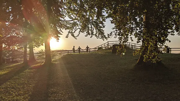 Eine stimmungsvolle Aufnahme bei Sonnenuntergang in einem Park oder auf einer Anhöhe. Die tief stehende Sonne strahlt hell links hinter einer Gruppe von Bäumen und erzeugt starke Lichtstrahlen und Silhouetten. Im Zentrum des Bildes stehen drei Personen Hand in Hand als Scherenschnitt vor dem hellen Horizont. Sie befinden sich auf einer erhöhten Grasfläche, die von einem rustikalen Holzzaun begrenzt wird. Der Vordergrund ist eine sanfte Grasböschung, die im Schatten liegt, während das Laub der Bäume im oberen Teil des Bildes vom Gegenlicht beleuchtet wird.