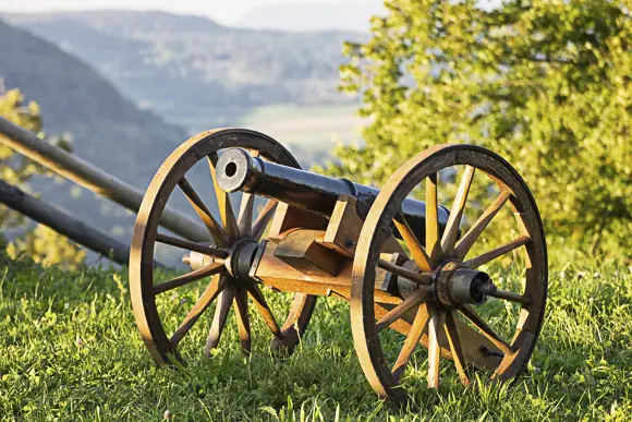 Eine historische Nachbildung einer kleinen Feldkanone oder einer Lafette auf einem grasbewachsenen Hügel, wahrscheinlich auf der Hiltenburg, bei Sonnenlicht. Die Kanone hat einen schwarzen Lauf und steht auf großen, hölzernen Speichenrädern und einer hölzernen Lafette. Im Hintergrund erstreckt sich eine weite, grüne, hügelige Landschaft oder ein bewaldetes Tal. Die Sonne beleuchtet die Kanone und einen hellgrünen Baum oder Busch rechts daneben.