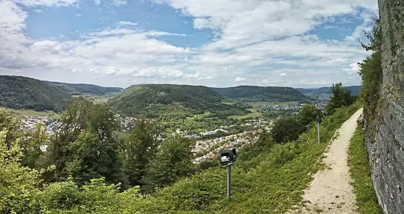 Ein weites Panorama, aufgenommen von einem erhöhten Standpunkt, wahrscheinlich der Hiltenburg, mit Blick über das Tal und die Ortschaft Bad Ditzenbach. Im Vordergrund verläuft ein schmaler, heller Wanderweg entlang einer historischen Steinmauer, umgeben von dichtem, grünem Gestrüpp und Bäumen. Der Blick fällt auf ein breites, bewaldetes Tal mit sanften, grünen Hügeln und Wäldern. In der Talsohle ist die lockere Bebauung einer Ortschaft (Bad Ditzenbach) zu erkennen. Über der Szene spannt sich ein blauer Himmel mit großen, weißen Cumuluswolken.