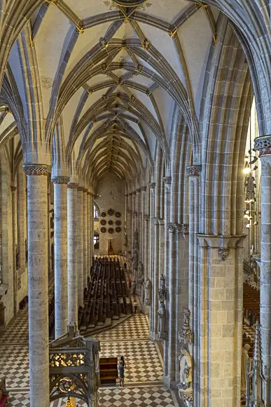 Eine hochwinkelige Innenansicht eines Münsters (des Ulmer Münsters), die das Seitenschiff und den Chor zeigt. Die Szene ist geprägt von hohen, schlanken, gerippten Säulen und Spitzbögen, die in ein Kreuzrippengewölbe münden. Der Boden hat ein markantes rot-weißes Schachbrettmuster. In der Tiefe des Raumes sind Reihen von dunklen Kirchenbänken oder Chorgestühlen zu sehen. Eine einzelne Person, klein im Vergleich zur Architektur, steht auf dem Boden. Das Licht fällt von oben und beleuchtet die hellen Steinsäulen und das Gewölbe.