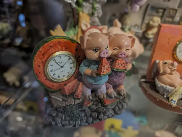 Eine detailreiche, kitschige Dekorationsfigur aus Harz oder Keramik, die zwei kleine, lächelnde Schweinchen beim Meloneessen darstellt. Die Schweinchen stehen vor einer Uhr, deren Zifferblatt in die Form einer aufgeschnittenen Wassermelone integriert ist. Im Hintergrund sind weitere Schweinefiguren unscharf zu erkennen.