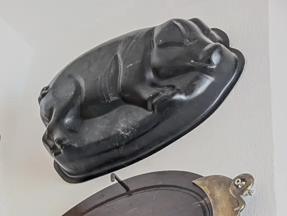 Eine historische, dunkle Kuchen- oder Pastetenform aus Metall, die ein liegendes Schwein dreidimensional darstellt. Die Backform hängt schräg an einer weißen Wand, darunter ist ein Teil eines anderen runden, dunklen Dekorationsgegenstandes mit einem metallenen Detail zu sehen.