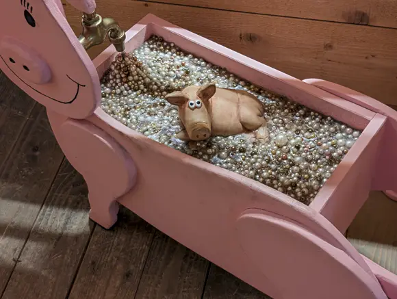 Eine humorvolle Installation: Eine rosa, schwein-förmige Holzbadewanne, gefüllt mit vielen glänzenden Perlen. In der Mitte der Perlen liegt eine kleine Keramik-Schweinefigur mit großen, runden Augen. Aus einem Wasserhahn oberhalb des Schweins scheint eine Perlenschüttung das Badewasser zu imitieren.