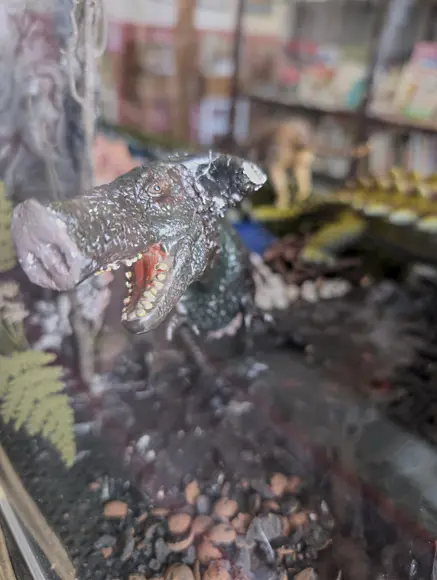 Eine Nahaufnahme einer Diorama-Figur in dunklen Farben, die einen prähistorischen, schweinähnlichen Dinosaurier oder ein Ur-Schwein (möglicherweise ein Entelodont) mit weit geöffnetem Maul und sichtbaren Zähnen darstellt. Die Figur ist in einer dunklen, erdigen Umgebung platziert, mit einem Farnblatt im Vordergrund.