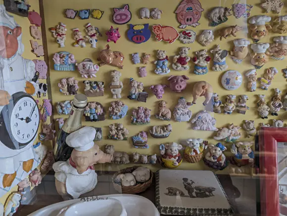 Eine gelbe Wand, die dicht mit einer Vielzahl kleiner Schweinefiguren in verschiedenen Formen, Farben und Posen bedeckt ist. Die dreidimensionalen Exponate zeigen Schweine als Köche, in traditioneller Kleidung, als Babys oder als Comicfiguren. Im unteren linken Bereich steht eine große Koch-Schweine-Figur neben einer Flasche Sekt und einer Wanduhr.