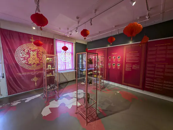 Ein Ausstellungsraum in einem Schweinemuseum, der dem chinesischen Jahr des Schweins gewidmet ist. Die Wände sind mit leuchtend rotem Stoff und Bannern mit goldenen Mustern und chinesischen Laternen dekoriert. In der Mitte des Raumes stehen verglaste Vitrinen mit Exponaten. Auf dem Boden ist eine stilisierte Weltkarte in Grau- und Rottönen abgebildet.