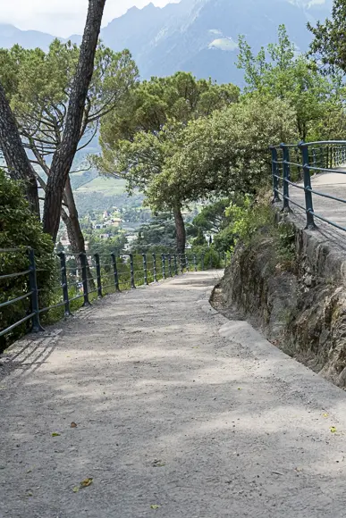 Ein Schotterweg, gesäumt von einem schwarzen Metallgeländer und einigen hohen Bäumen (vermutlich Kiefern), führt bergab an einer Felsmauer vorbei. Die Szene ist sonnig und eröffnet in der Ferne einen weiten Blick auf eine von Wald umgebene Tallandschaft und im Hintergrund hohe Berge in Meran, Südtirol.