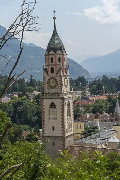 Ein imposanter, historischer Kirchturm der Stadtpfarrkirche St. Nikolaus in Meran mit einer schlanken, grünen Spitze und mehreren Uhrwerken dominiert das Bild. Der Turm hat eine helle, verzierte Fassade mit rosa Akzenten und hohen Bogenfenstern. Er ragt über eine dicht bebaute Stadtlandschaft mit roten Dächern und vielen Bäumen empor. Im Hintergrund erheben sich majestätische Berge unter einem leicht bewölkten Himmel. Im Vordergrund sind Äste und grüne Blätter zu sehen, die einen Rahmen bilden.