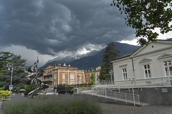 Eine dramatische Wolkenstimmung mit dunklen, tiefhängenden Gewitterwolken liegt über der Stadt Meran und den hohen Bergen im Hintergrund. Im Vordergrund rechts steht ein helles, klassizistisches Gebäude mit Rampe und Geländer. In der Mitte sind ältere Stadthäuser in warmen Farben wie Terrakotta und Gelb zu sehen. Links steht eine moderne, silberne Skulptur in Spiralform. Der Platz ist mit Pflastersteinen und Pflanzen, darunter Lavendel, gestaltet.