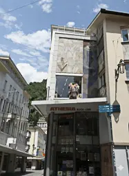 Ein kleines, buntes Kuscheltier sitzt auf einem Vordach über dem Eingang eines Geschäfts namens "ATHESIA BUCH-LIBRI" in einer sonnigen Straße in Meran. Das Gebäude hat eine helle Fassade mit kunstvollen Mosaiken und Erkerfenstern. Im Hintergrund sind weitere Gebäude und ein grüner Hügel unter einem blauen Himmel mit einigen Wolken zu sehen.