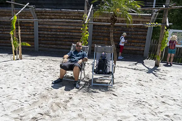 Christopher sitzt in einem Liegestuhl auf einem Sandboden, der einen künstlichen Strand im Freien nachbildet. Er trägt ein kariertes Hemd und kurze Hosen. Neben ihm steht eine kleine Palme und ein zweiter, leerer Campingstuhl mit einem Rucksack darauf sitze ich Klein Viki. Im Hintergrund befindet sich eine rustikale Holzwand.