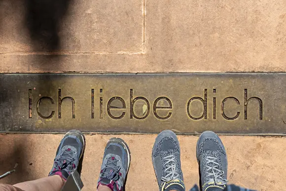 Aufnahme von oben, die eine Metallplatte mit der lasergravierten Aufschrift „Ich liebe dich“ zeigt, die in einen hellbraunen Pflasterboden eingelassen ist. Man sieht die Füße von zwei Personen in Wanderschuhen oder Turnschuhen, die jeweils links und rechts der Platte stehen.