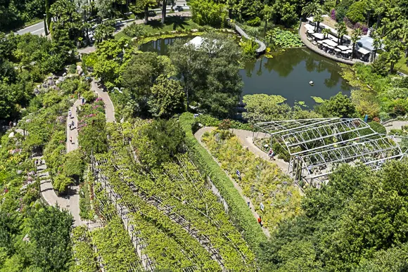 Blick von oben auf die Gärten von Schloss Trauttmansdorff mit Weinbergen, Gewächshäusern und einem Teich mit Restaurant.