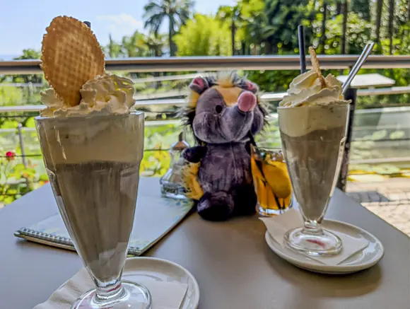 Zwei Eiskaffees und das Plüschtier "klein Viki" auf einem Tisch vor einem grünen, sommerlichen Hintergrund.