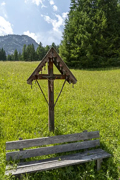 Ein traditioneller, überdachter hölzerner Bildstock mit einem Kruzifix steht inmitten einer weiten, sonnigen grünen Almwiese. Davor befindet sich eine einfache, rustikale Holzbank. Im Hintergrund sind ein dunkler Wald und ein bewaldeter Berg zu sehen.