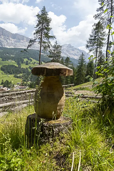Eine große, geschnitzte Holzskulptur in Form eines Pilzes (vermutlich eines Steinpilzes) steht auf einem Baumstumpf inmitten von hohem Gras. Im Hintergrund erstreckt sich eine grüne alpine Landschaft mit einem Tal und einem beeindruckenden, felsigen Bergmassiv unter einem bewölkten Himmel.
