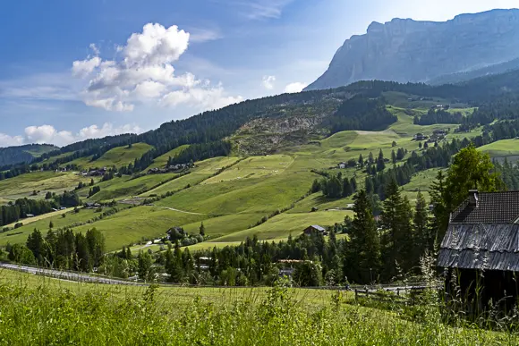 Eine weite Landschaft in Südtirol mit sanften grünen Hügeln, die mit kleinen Häusern und Wäldern bedeckt sind, unter einem teilweise bewölkten blauen Himmel. Im Hintergrund erheben sich hohe, felsige Berge.