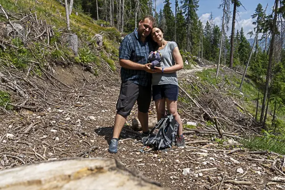 Mama und Papa stehen eng beieinander, sich umarmend, auf einem unbefestigten Waldweg in Alta Badia. Papa, mit Bart und kariertem Hemd, hält zusammen mit Mama, die ein graues Oberteil und eine kurze Hose trägt, das Kuscheltier von Klein Viki in ihren Händen. Im Hintergrund sind gefällte Baumstämme und ein Nadelwald zu sehen.
