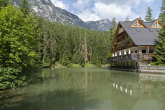 Ein großes, traditionelles Holzhaus mit Satteldach und einem langen Balkon mit weißen Schirmen, das direkt am Ufer eines ruhigen, waldgesäumten Sees steht. Im Hintergrund erheben sich steile, felsige Berge unter einem teilweise bewölkten Himmel.