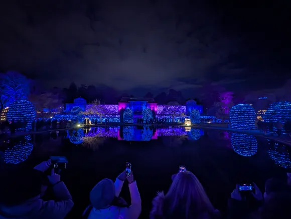 Silhouetten von Besuchern im Vordergrund, die mit ihren Smartphones das blau beleuchtete Schloss und die Lichtinstallationen fotografieren.