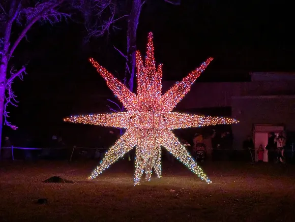 Eine große, aufrecht stehende Lichtskulptur in Sternform aus warmweißem Licht, platziert auf einer Wiese neben einem befestigten Weg.