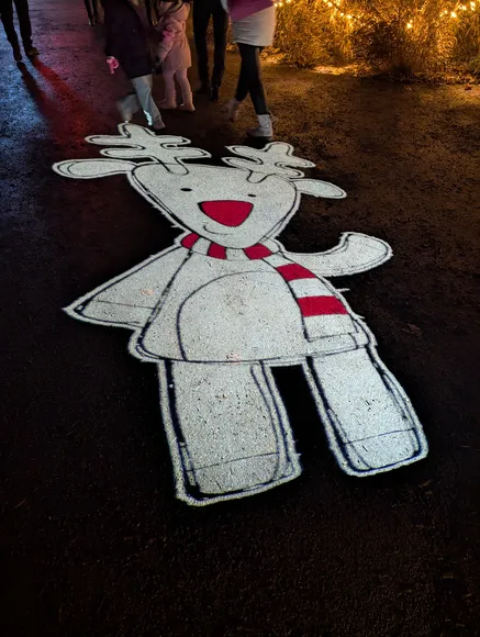 Eine Lichtprojektion auf dunklem Asphalt zeigt ein weißes Comic-Rentier mit roter Nase und rot-weiß gestreiftem Schal.
