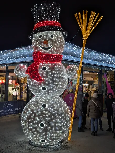 Eine mehrstöckige, riesige Schneemann-Lichtfigur mit schwarzem Zylinder, rotem Licht-Schal und einem gelben Licht-Besen vor einem hell erleuchteten Gebäude.